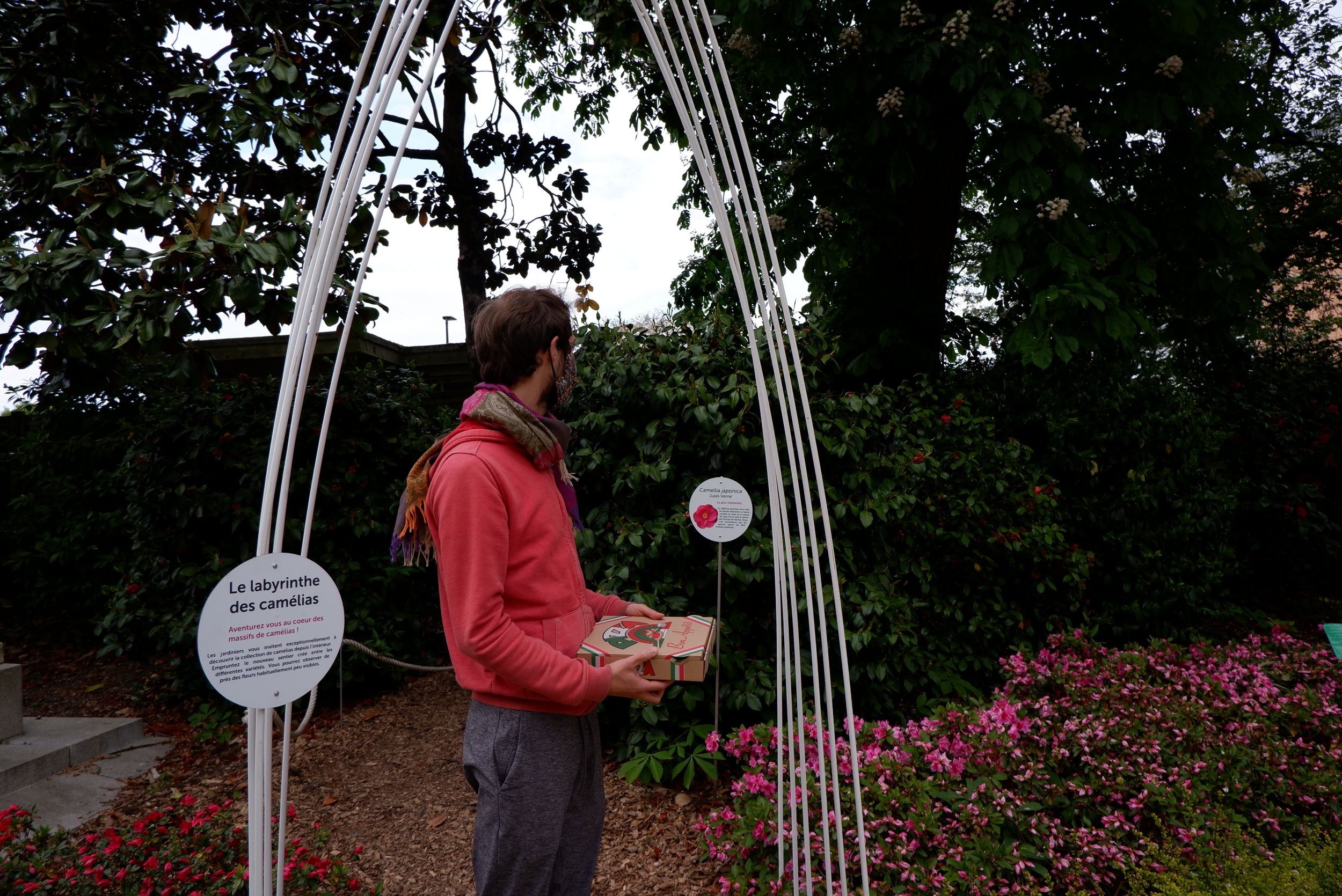 Ryan portant une pizza en face du labyrinthe des camélias dans le jardin des plantes