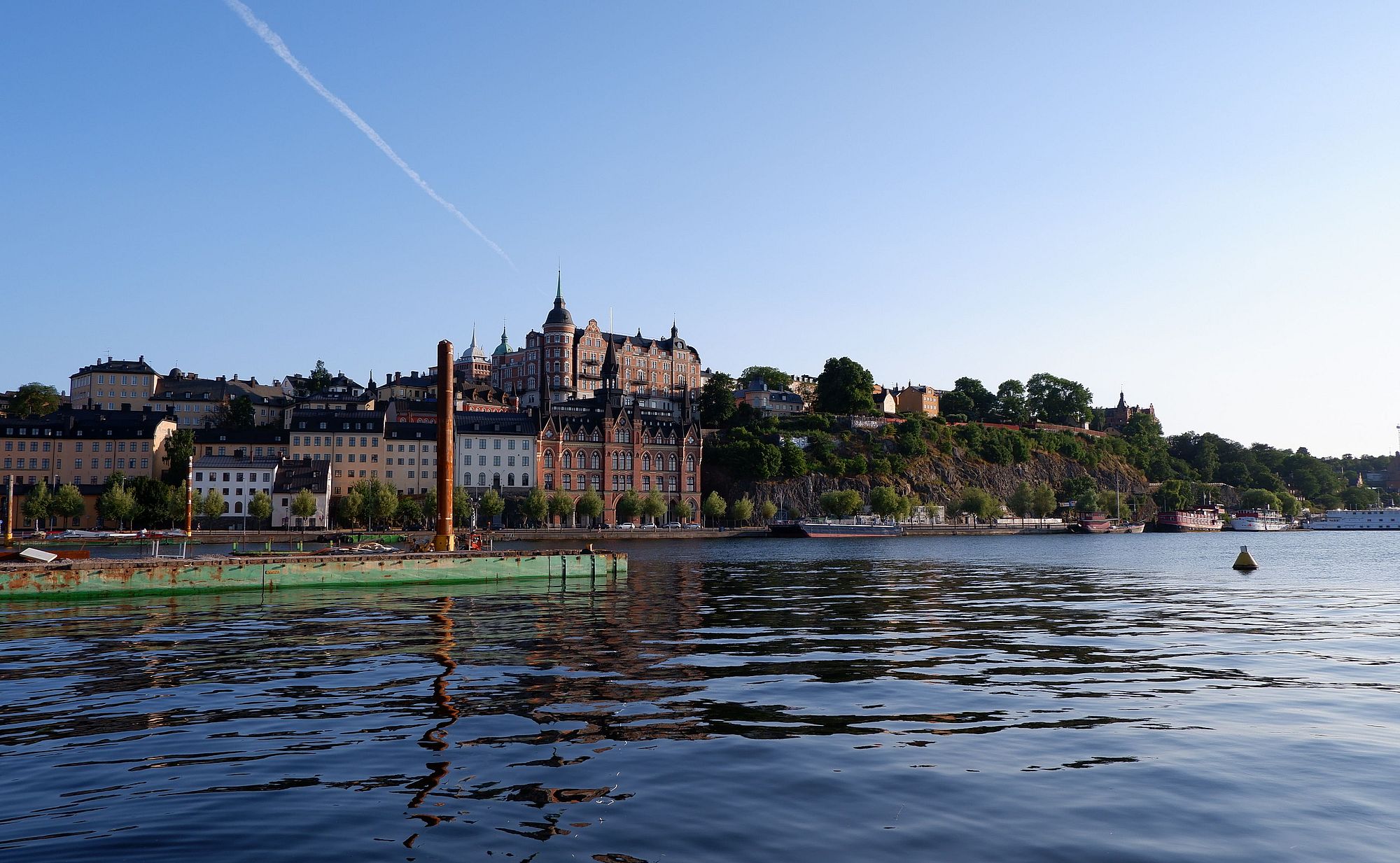 Un paysage représentant Stockholm avec une grande étendue d'eau au premier plan et des bâtiments pittoresques en arrière-plan, quasiment perchés sur la falaise.